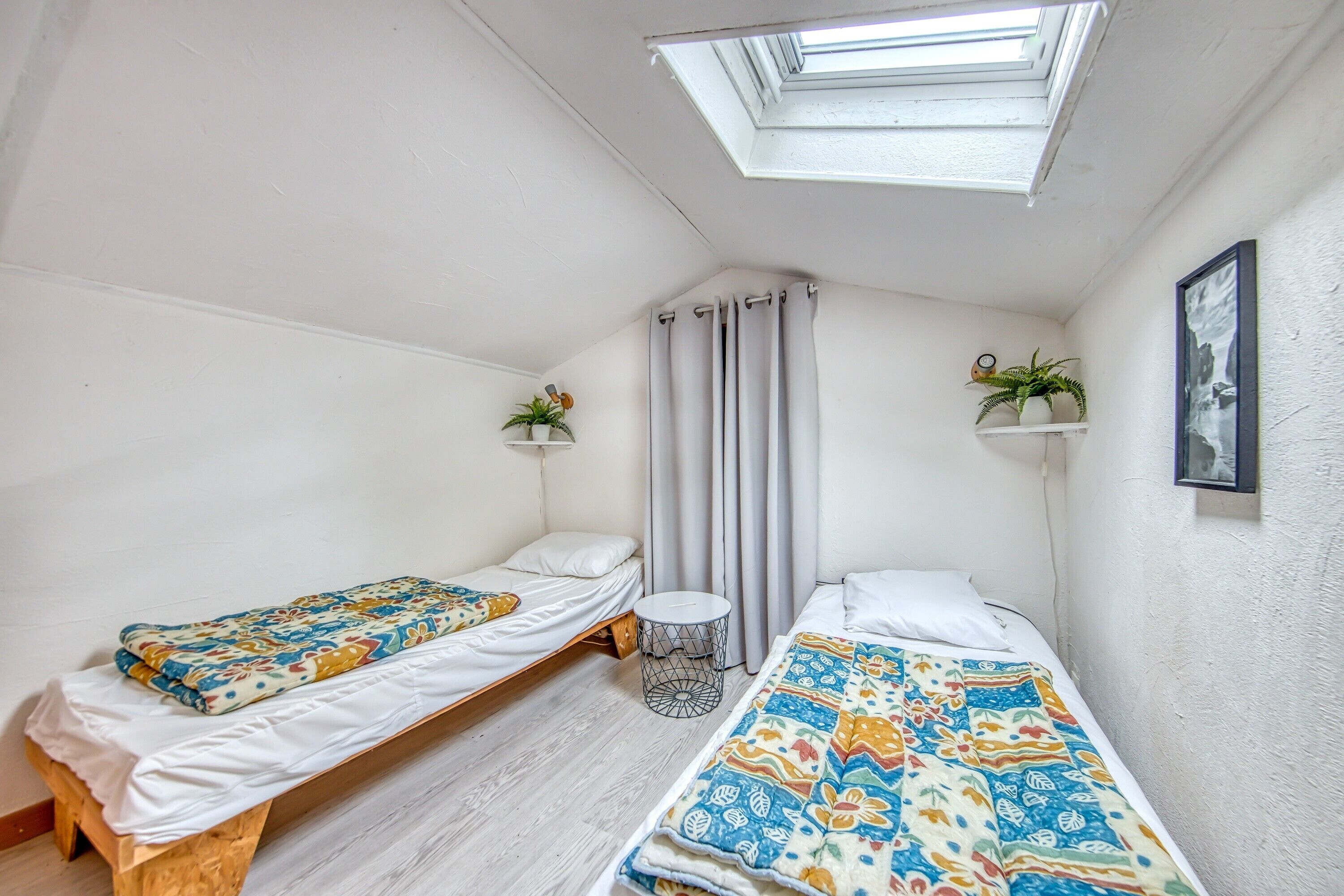 Una habitación pequeña y luminosa con dos camas individuales, un tragaluz, una mesita redonda y dos estantes con plantas.