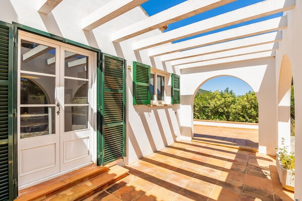A covered outdoor area with a white wall, green shutters, and a view of greenery.