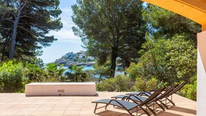 A patio with lounge chairs, a white planter, and a view of a coastal area with trees and a building on a hill. - Holiday Home 'Aladern' with Sea View, Private Terrace and Wi-Fi (Font de Sa Cala)