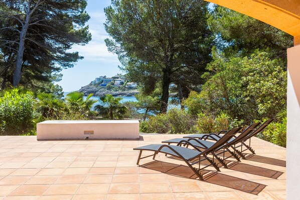 A patio with lounge chairs, a white planter, and a view of a coastal area with trees and a building on a hill. - Holiday Home 'Aladern' with Sea View, Private Terrace and Wi-Fi (Font de Sa Cala)