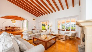 A spacious living room with a wooden ceiling, a fireplace, and a large window offering a view of greenery. - Holiday Home 'Aladern' with Sea View, Private Terrace and Wi-Fi (Font de Sa Cala)