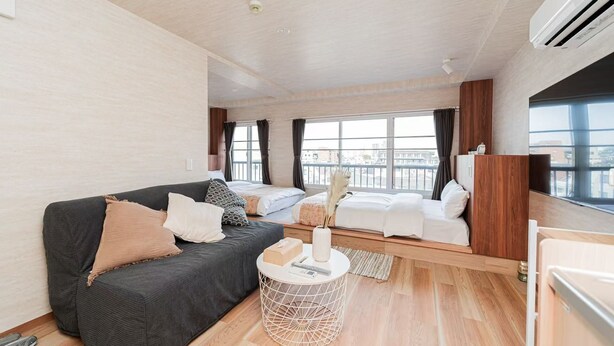 A modern hotel room with a grey sofa, a bed with white bedding, a small round table, and a view of the city through large windows.