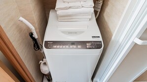 A compact, white, front-loading washing machine with a control panel, stacked with white towels, in a small bathroom with beige tiles.