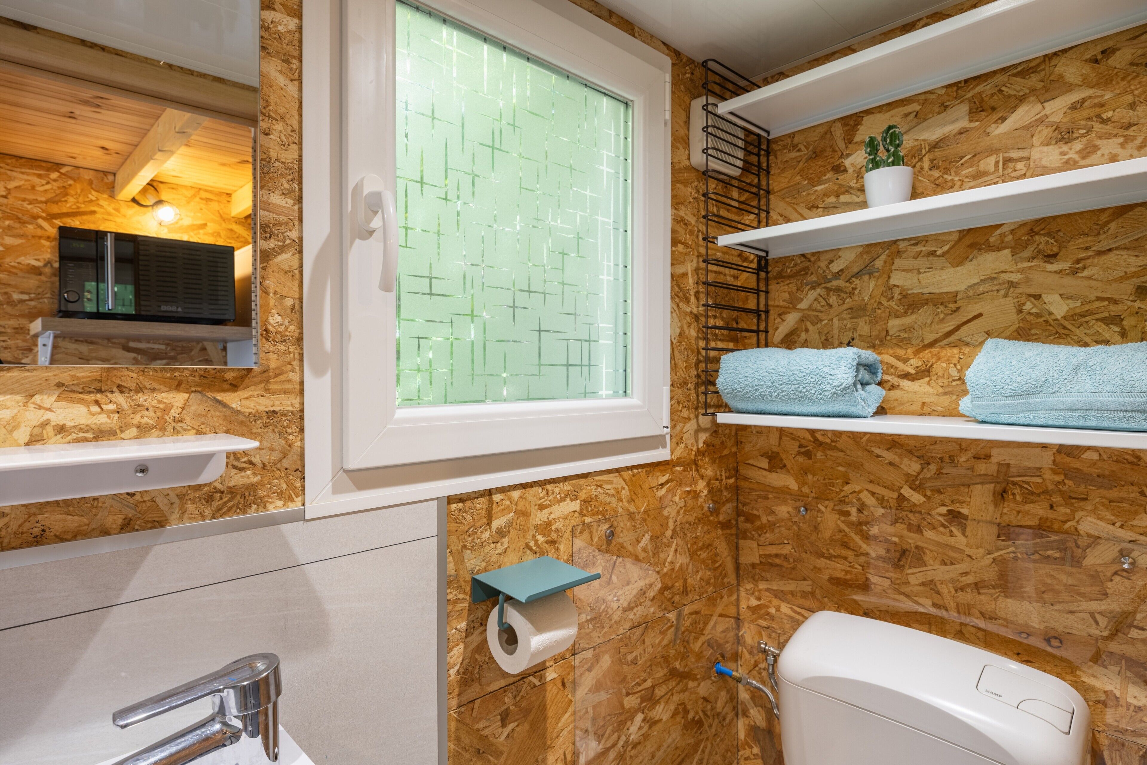 Een badkamer met een houten wand, een raam met een geometrisch patroon, een toilet, een wastafel en een plank met handdoeken.