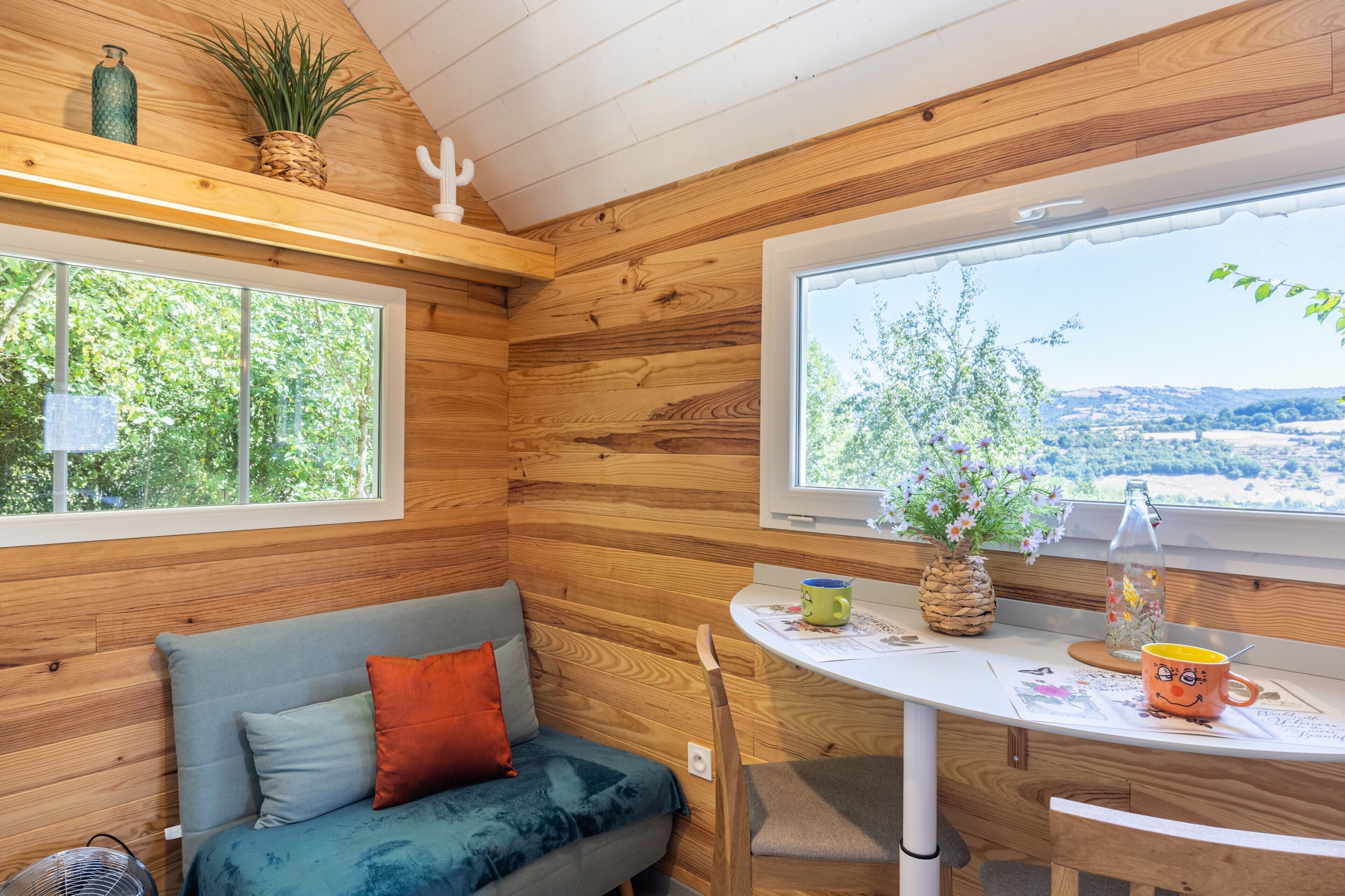 A cozy cabin with wooden walls, a cushioned bench, a table with a mug, and a window with a view of greenery.