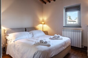 Un lit bien fait, avec du linge de lit blanc, une lampe sur une table de chevet et une fenêtre donnant sur les bâtiments environnants.