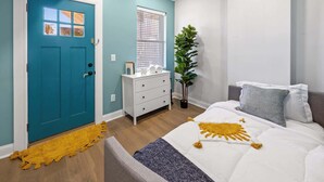 Une chambre avec une porte de couleur turquoise, une commode blanche, un lit avec une literie blanche et grise, et une plante.