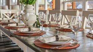 Une table à manger dressée avec des assiettes, des couverts et des verres à vin, agrémentée d’une plante en pot et offrant une vue sur l’extérieur grâce à de grandes fenêtres.