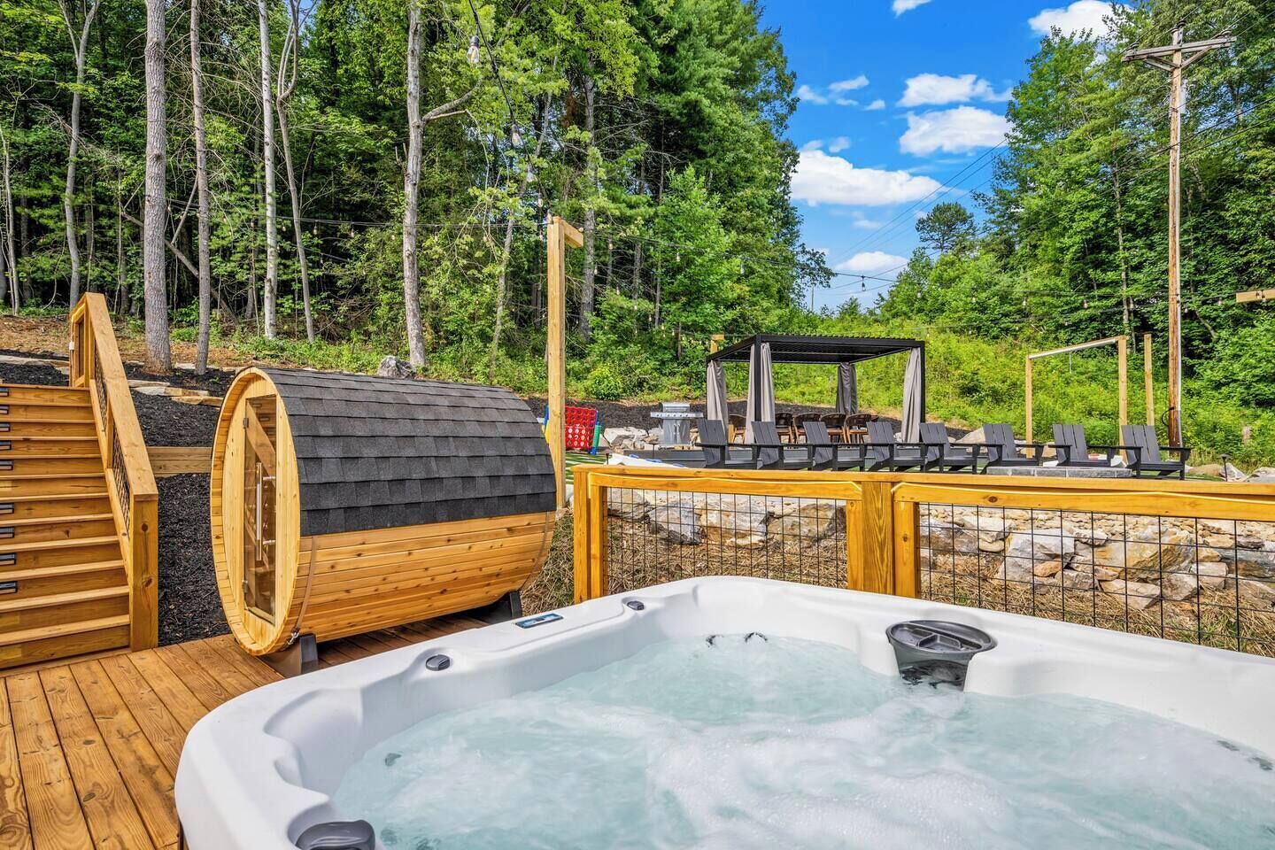 Ein Whirlpool auf einer Holzterrasse mit Sauna und Sitzgelegenheit inmitten eines bewaldeten Gebietes.