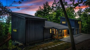 A modern two-story house with a dark exterior, a wooden deck, and a visible staircase leading to the entrance.