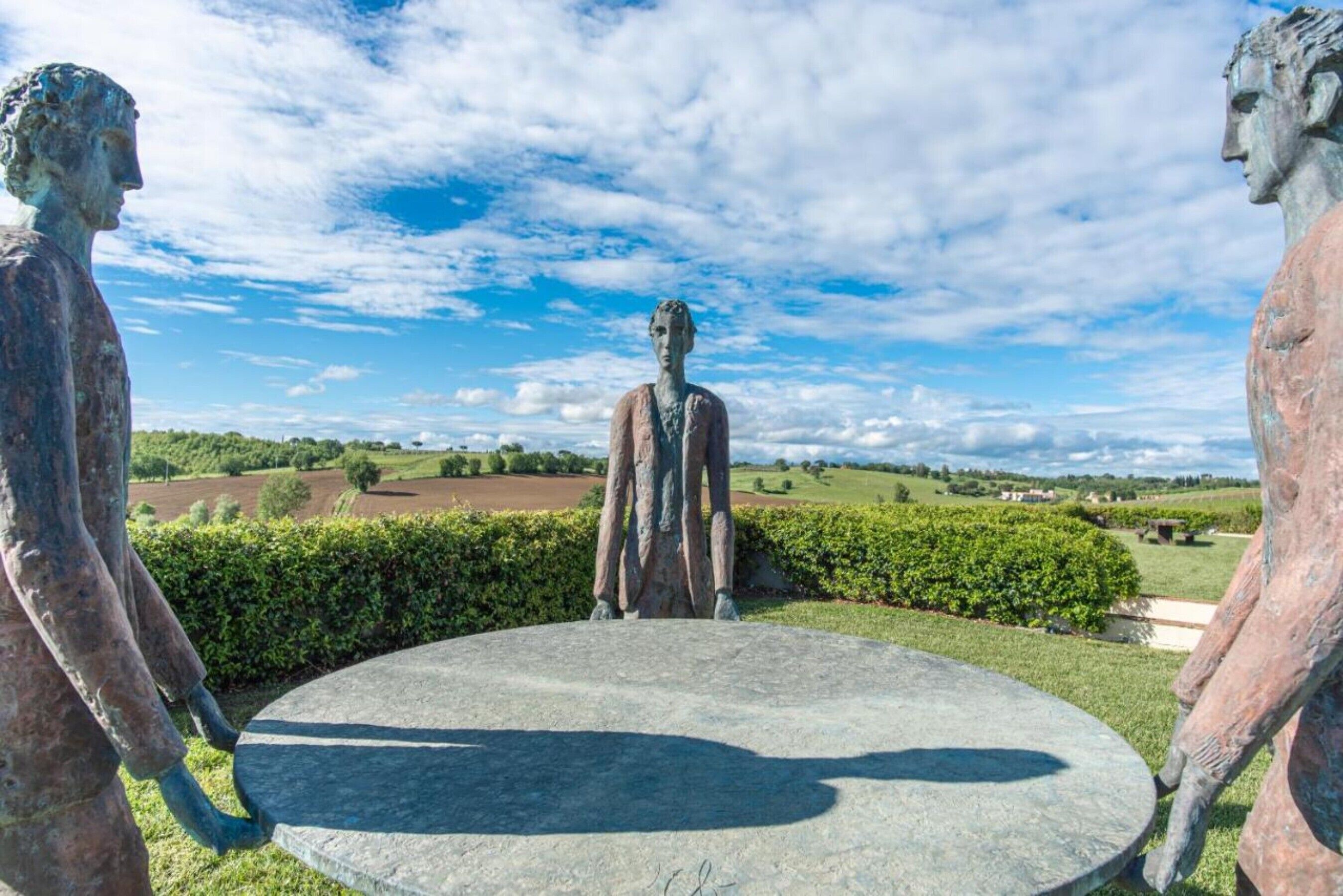 Una statua con un tavolo di pietra circolare di fronte, situata in un'area paesaggistica con siepi e campi aperti.