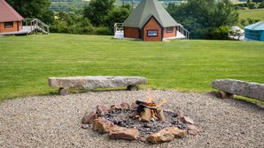 A lodge with a fire pit in the foreground, surrounded by a grassy field and a view of rolling hills in the background. - Dartmoor Eco-Pod (Oak Tree Lane) Gorgeous rustic charm (Okehampton)