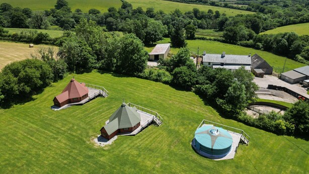 An aerial view of a rural property with multiple buildings, including a farmhouse, a barn, and a large circular structure, surrounded by lush green fields and trees. - Dartmoor Eco-Pod (Oak Tree Lane) Gorgeous rustic charm (Okehampton)