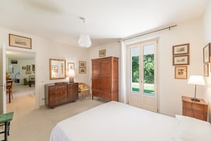 A bedroom with a bed, a wooden dresser, a chair, and a large window with a view of greenery.