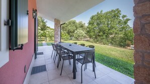 An outdoor patio with a table and chairs, a stone wall, and a view of a grassy area with trees. - ApartmentsGarda - Cottage Capre (GARDA)