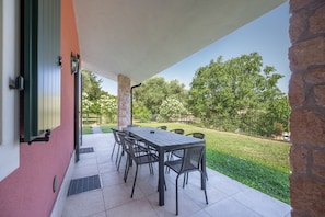 An outdoor patio with a table and chairs, a stone wall, and a view of a grassy area with trees. - ApartmentsGarda - Cottage Capre (GARDA)