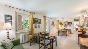 A spacious living area with a green sofa, wooden chairs, a dining table, and a kitchen in the background. - ApartmentsGarda - Cottage Capre (GARDA)