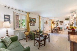 A spacious living area with a green sofa, wooden chairs, a dining table, and a kitchen in the background.