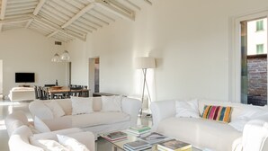 A spacious living area with a white sofa, a coffee table with books, a TV, and a dining area in the background. - Villa 'Loft' with Shared Pool, Wi-Fi and Air Conditioning (Cortona)