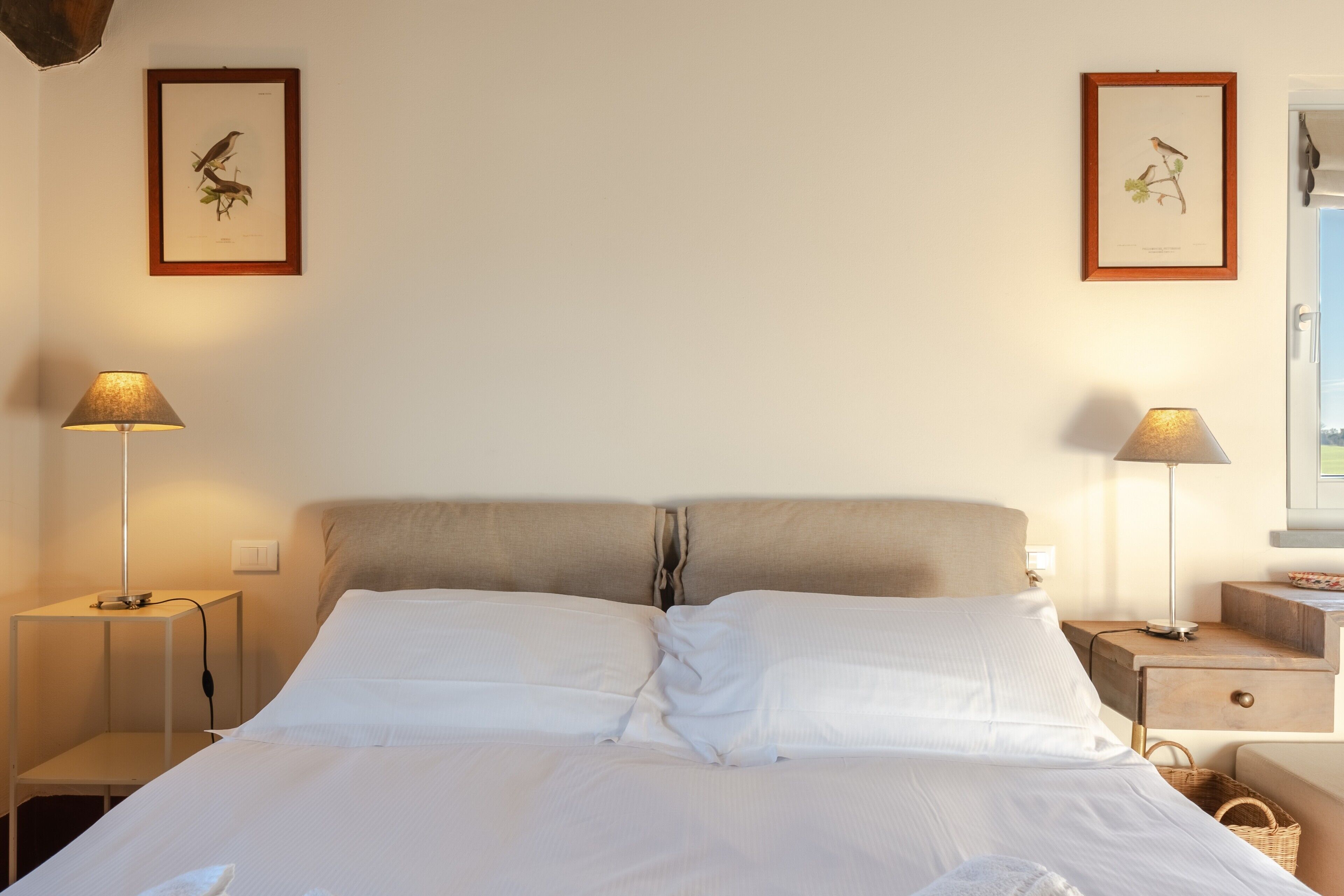 A neatly made bed with white linens, two bedside tables with lamps, and framed botanical prints on the wall.
