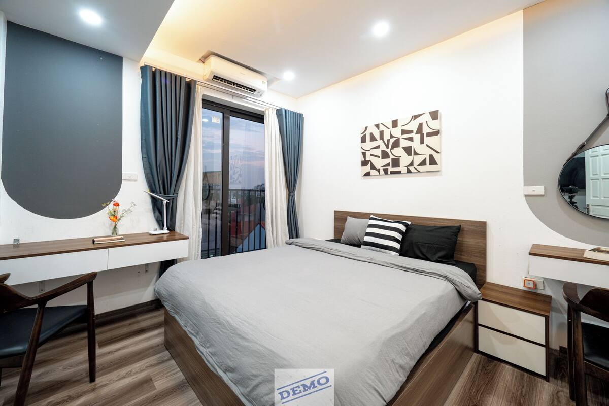 A modern hotel room with a bed, desk, and chair, a large window with curtains, and a wall-mounted air conditioner.