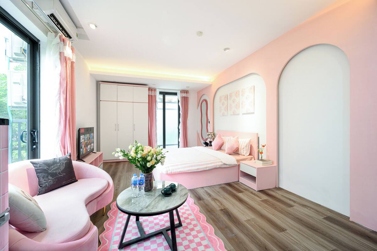 A pink-themed bedroom with a bed, a pink sofa, a small table with a vase of flowers, and a window with curtains.