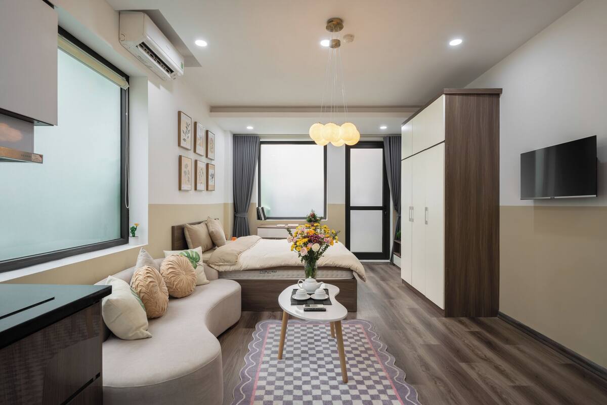 A modern hotel room with a sofa, a small table with a vase of flowers, a TV mounted on the wall, and a large window.