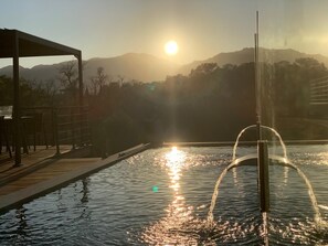 A pool with a fountain, surrounded by a wooden deck and a covered seating area, with mountains in the background.