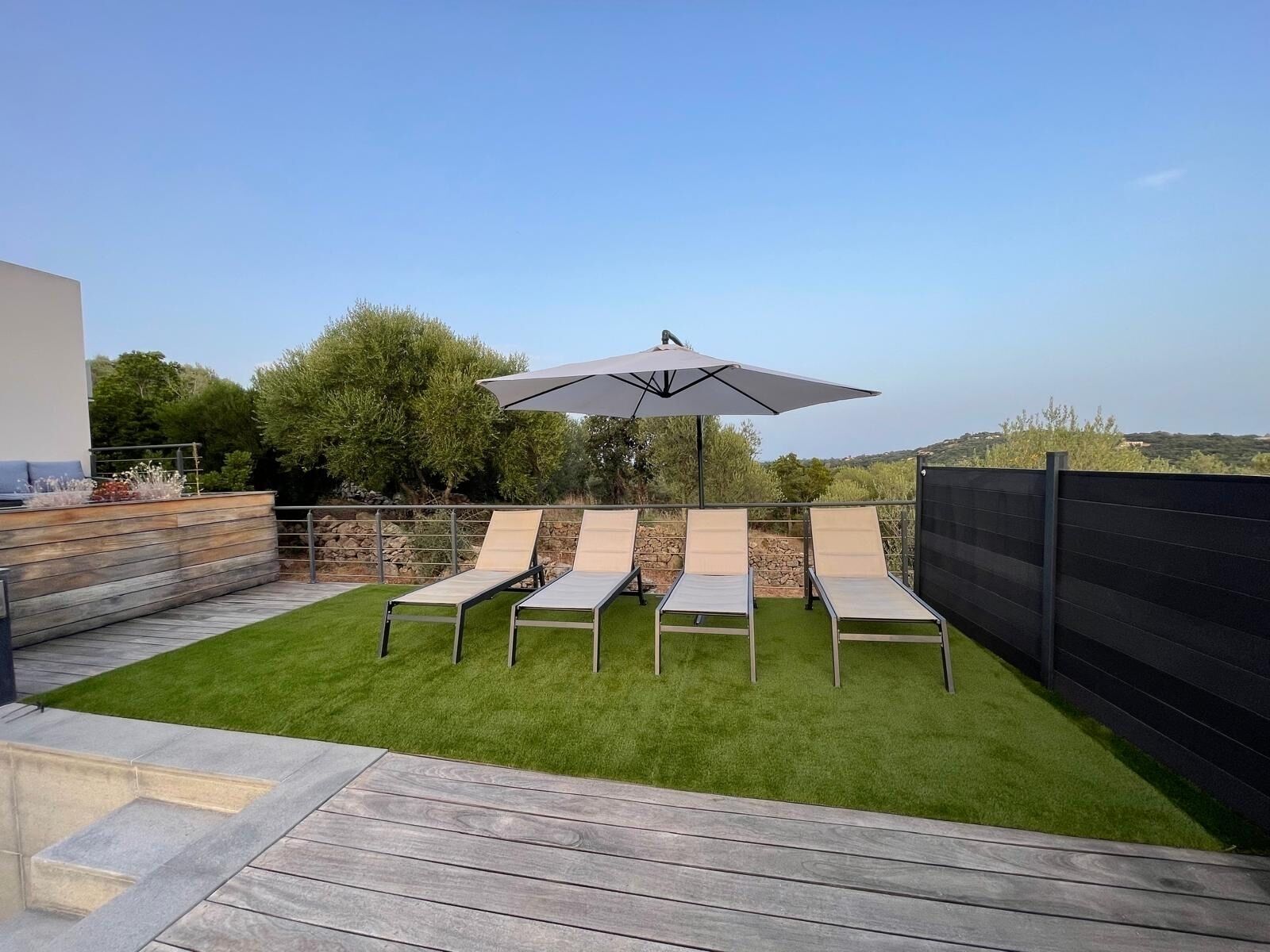 A patio with four lounge chairs and a large umbrella, surrounded by a wooden deck and a stone wall.