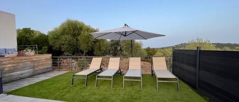 A patio with four lounge chairs and a large umbrella, surrounded by a wooden deck and a stone wall.