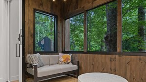Una habitación con piso de madera, un sofá con cojines estampados, una mesa redonda y ventanas amplias que dan a un paisaje de árboles.
