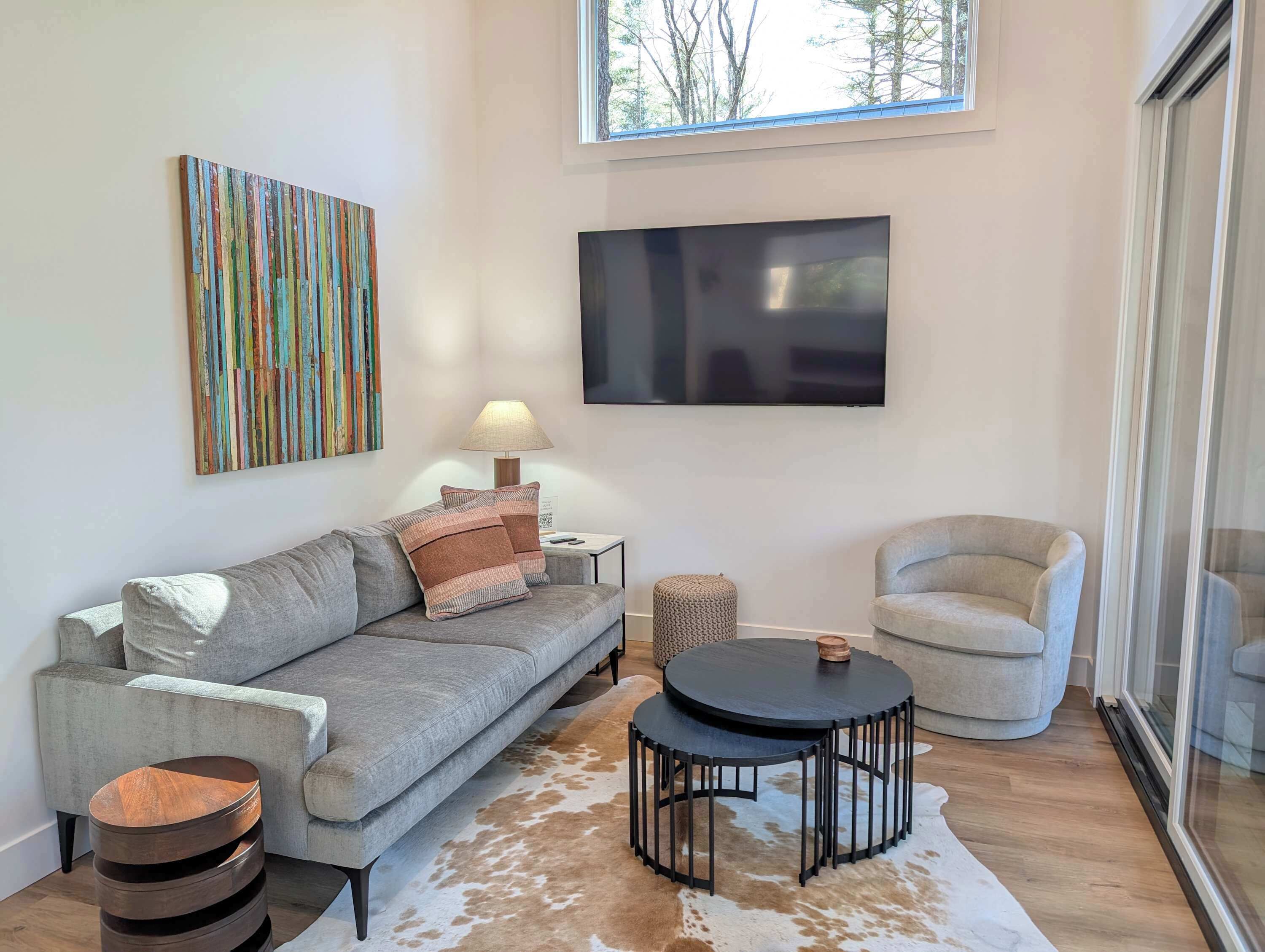 A modern living room with a grey sofa, a round coffee table, a TV mounted on the wall, and a window providing natural light.