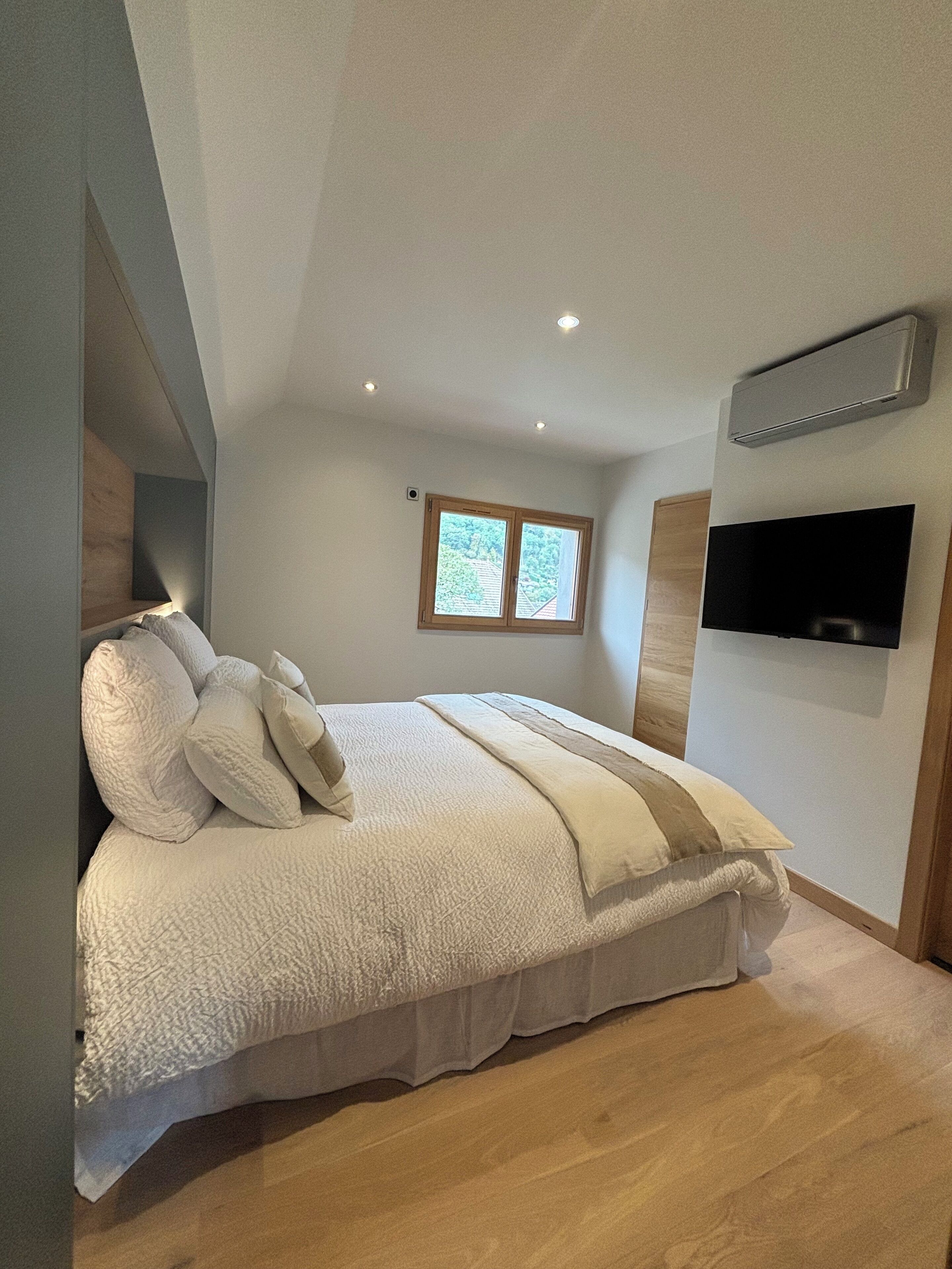 A bedroom with a large bed, a window, a flat-screen TV, and a ceiling fan.