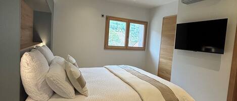 A bedroom with a large bed, a window, a flat-screen TV, and a ceiling fan.