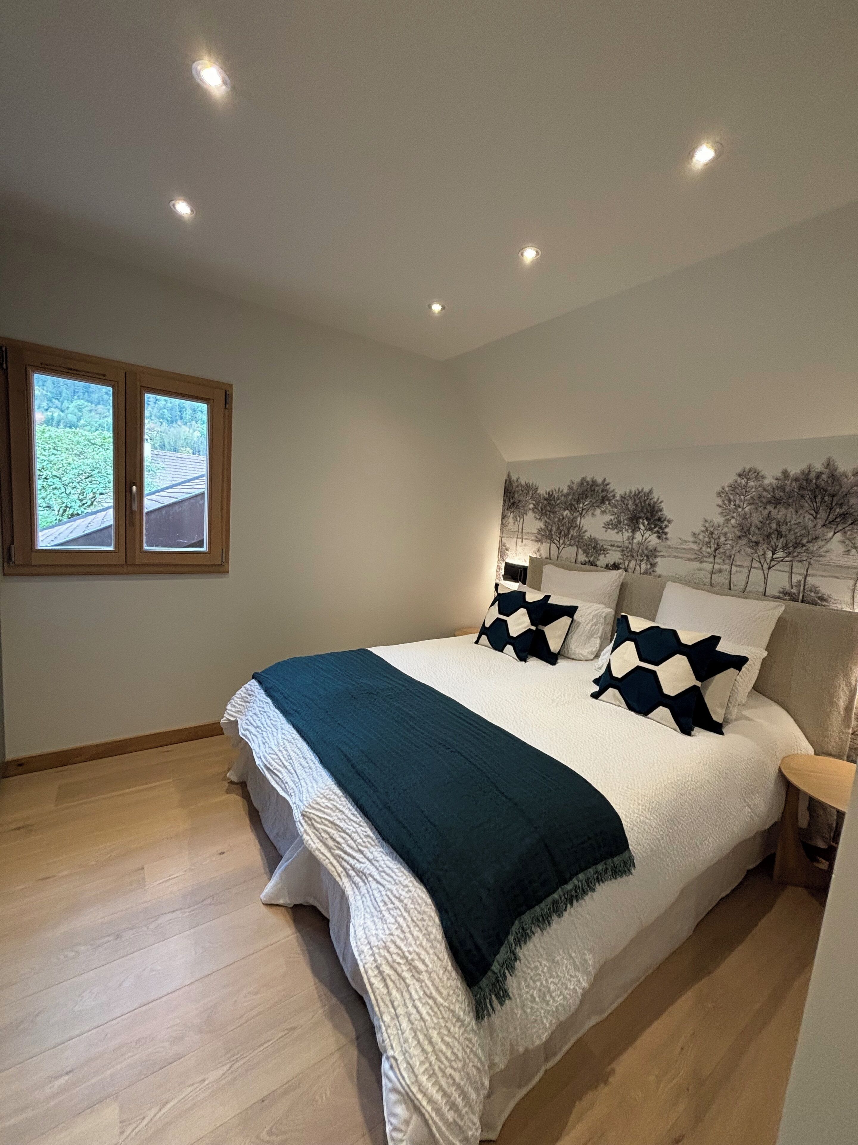 A bedroom with a large bed, a window with a view of greenery, and a ceiling with recessed lighting.
