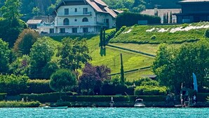 A large, multi-story house with a sloped roof, surrounded by lush greenery and a body of water. - Modern duplex with panoramic lake view & 100m from cycle path (Sévrier)