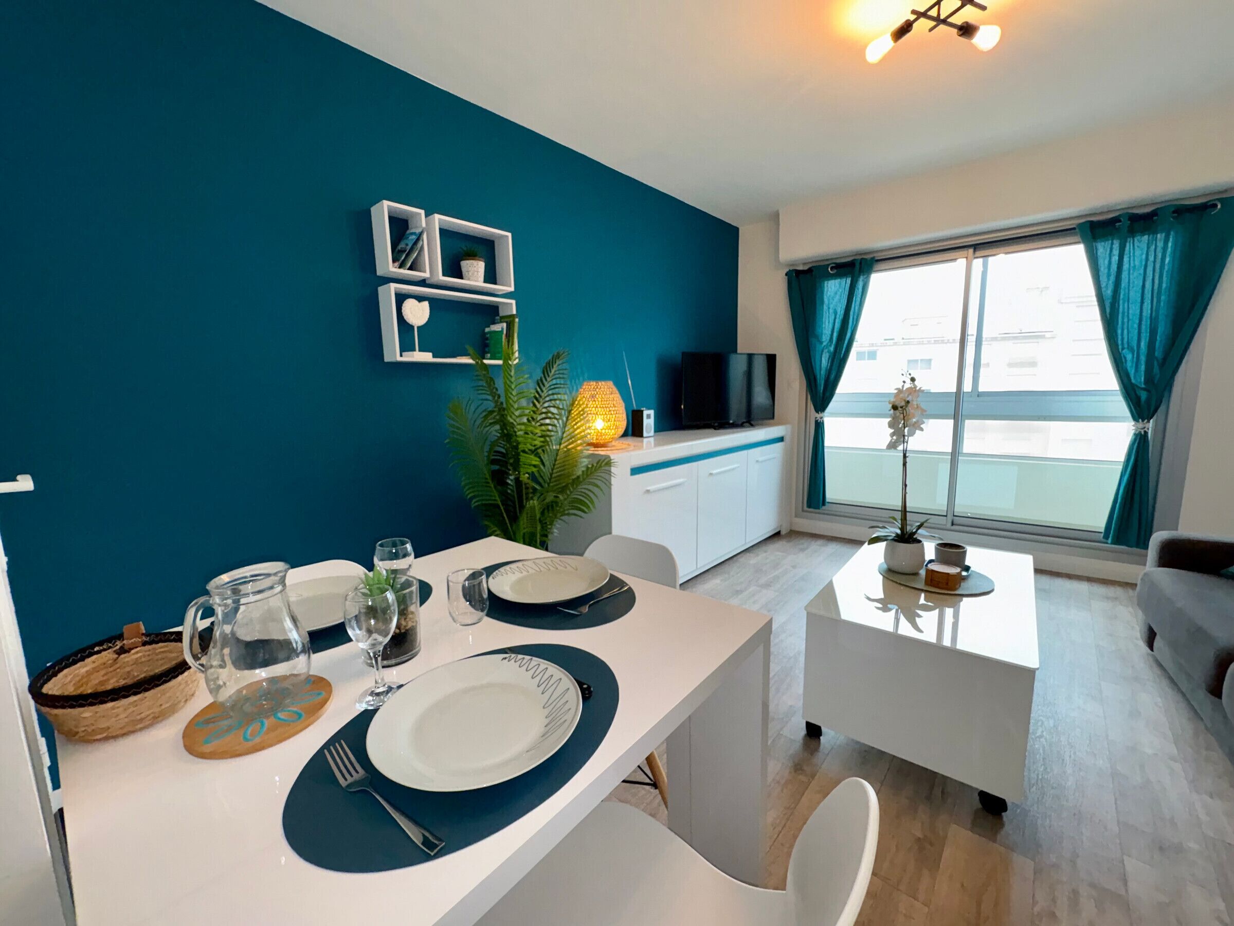 Uma sala de jantar moderna com uma mesa branca posta para dois, uma televisão em um armário branco, e uma janela grande com cortinas verde-água.