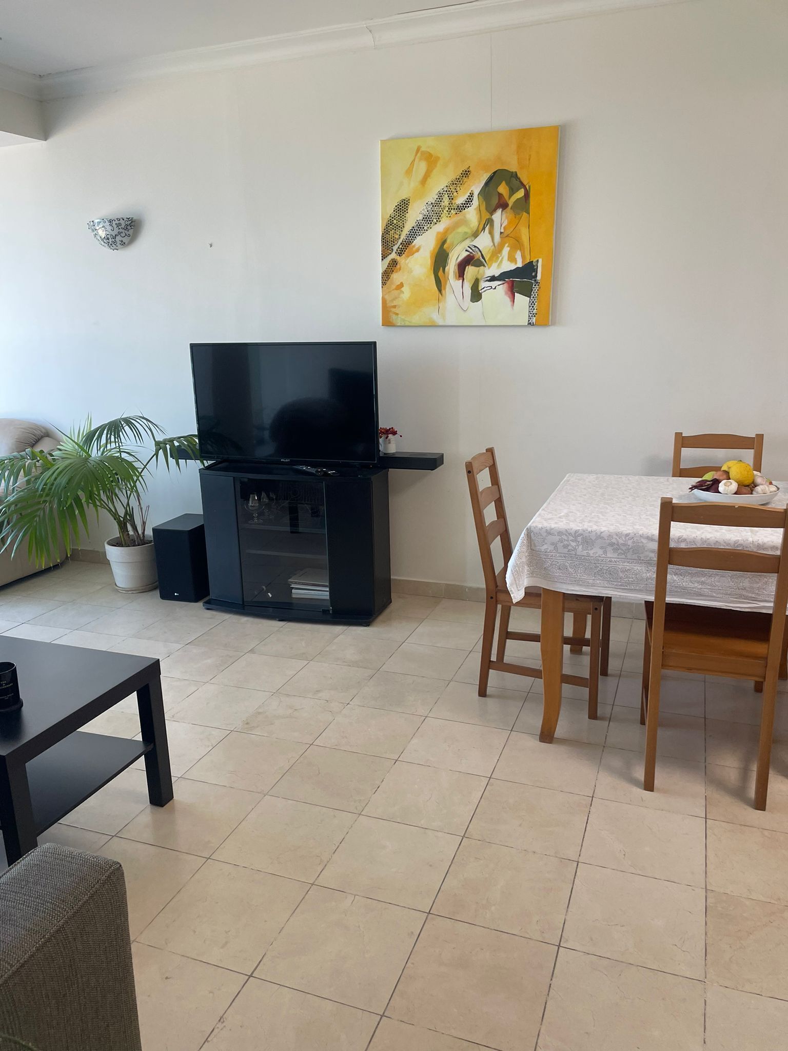 A living room with a flat-screen TV, a dining table with chairs, a potted plant, and a painting on the wall.