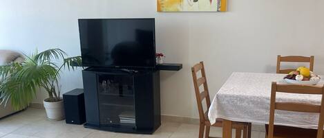 A living room with a flat-screen TV, a dining table with chairs, a potted plant, and a painting on the wall.