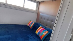 A single bed with a blue bedspread and two colorful pillows, a window with blinds, and a white door. - Cozy apartment for 2 with WIFI, air conditioning and parking in Cap d'Agde (Cap d'Agde naturisme)