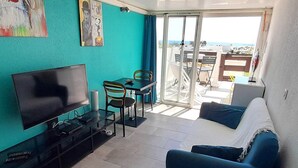 A living room with a TV, a sofa, a dining table with chairs, and a balcony with a view. - Cozy apartment for 2 with WIFI, air conditioning and parking in Cap d'Agde (Cap d'Agde naturisme)