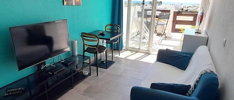 A living room with a TV, a sofa, a dining table with chairs, and a balcony with a view.