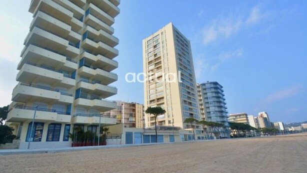 Un hôtel en bord de mer, composé de bâtiments à plusieurs étages, avec une plage de sable en premier plan et un ciel dégagé.