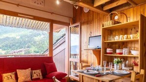 A cozy wooden cabin with a red sofa, dining table, and chairs. A TV is mounted on the wall, and there are shelves with kitchenware. - Alpine mezzanine studio, sleeps 4, pets allowed, optional parking (Valmorel)