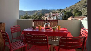 A balcony with a red table and chairs, a kettle, and a view of a hillside town.