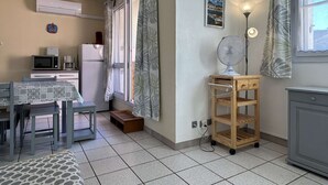 A compact kitchen with a microwave, coffee maker, and refrigerator. There is a dining table with chairs, a small wooden cabinet, and a fan on a cart.