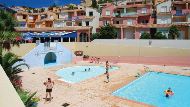 A swimming pool area with multiple pools, a covered seating area, and a view of residential buildings in the background.