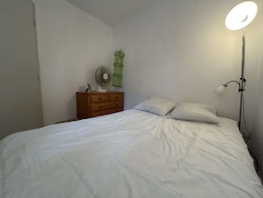 A bedroom with a bed, a dresser, a fan, and a bedside lamp. - Cozy apartment, 2 rooms, near beach with secure parking (Fleury-Saint-Pierre-la-Mer)