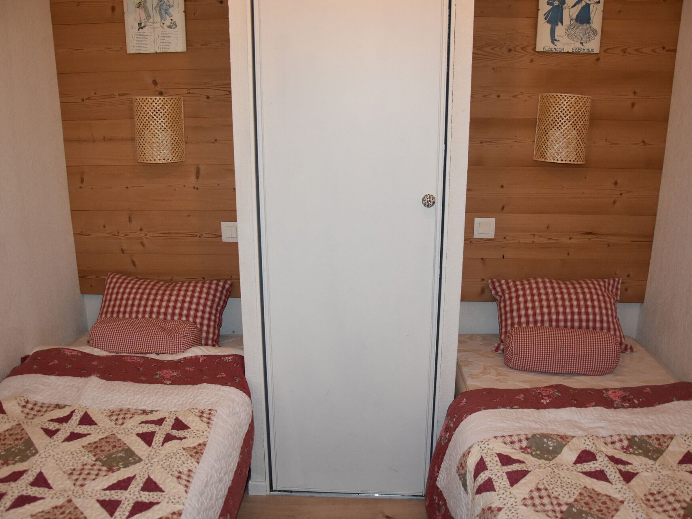 A room with two beds, each with a red and white checkered pillow and a quilt with a geometric pattern.