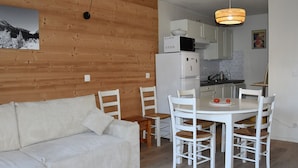 Un salon chaleureux avec un canapé blanc et une table à manger entourée de chaises. On aperçoit à l’arrière-plan un coin cuisine avec des meubles blancs et des appareils électroménagers.